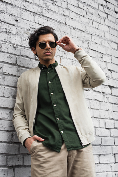 young man in stylish sunglasses and bomber jacket posing with hand in pocket near brick wall - Foto, immagini