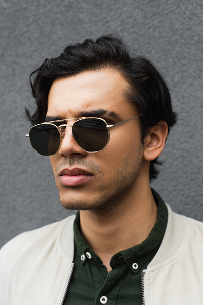 retrato de un joven con gafas de sol elegantes posando cerca de la pared gris - Foto, Imagen