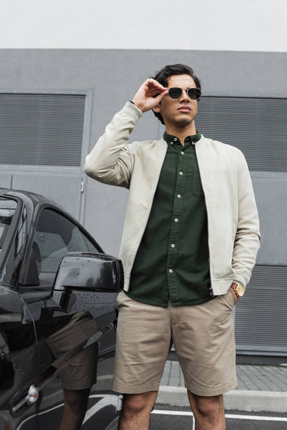 young man in stylish sunglasses and bomber jacket standing with hand in pocket near modern car outside - Foto, Imagem