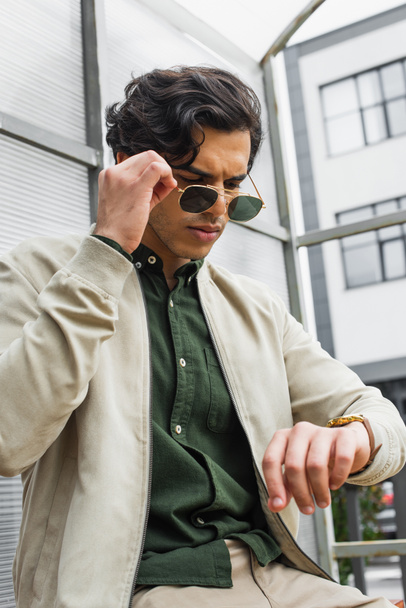 young man adjusting stylish sunglasses while looking at wristwatch  - Foto, Bild