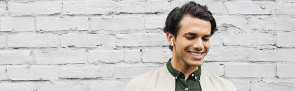 vidám fiatalember vezeték nélküli fülhallgató mosolyog, miközben zenét hallgat közelében téglafal, banner - Fotó, kép