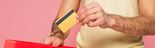 vista recortada del joven que pone la tarjeta de crédito en el bolso rojo de la compra aislado en rosa, bandera - Foto, imagen