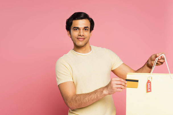 vrolijke jongeman met credit card en boodschappentas met sale tag geïsoleerd op roze - Foto, afbeelding