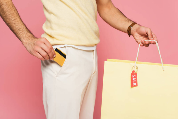 vista recortada de un hombre joven que pone la tarjeta de crédito en el bolsillo y la celebración de la bolsa de compras con etiqueta de venta aislado en rosa - Foto, imagen