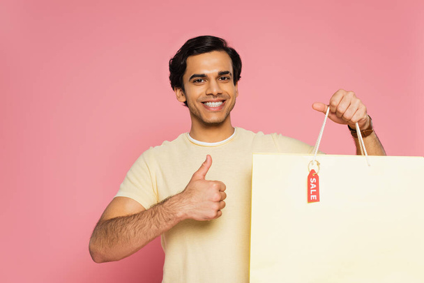 sorridente jovem segurando saco de compras com etiqueta de venda e mostrando polegar para cima isolado em rosa - Foto, Imagem