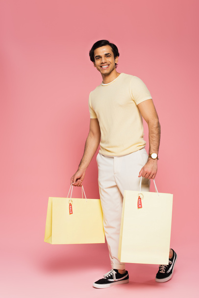 full length of cheerful young man holding shopping bags with sale tags on pink - Fotó, kép