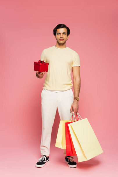 πλήρες μήκος του νεαρού άνδρα κρατώντας κόκκινο κουτί δώρου και τσάντες ψώνια σε ροζ - Φωτογραφία, εικόνα
