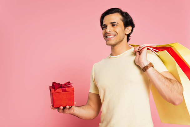 Lächelnder junger Mann mit roter Geschenkschachtel und Einkaufstaschen auf rosa - Foto, Bild