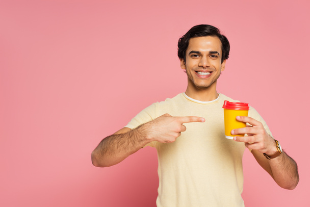 happy young man pointing with finger at paper cup with coffee to go isolated on pink - 写真・画像