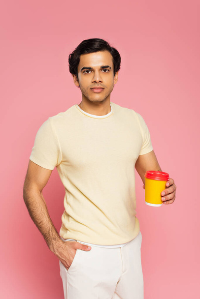 young man holding paper cup with coffee to go while standing with hand in pocket isolated on pink - Foto, imagen