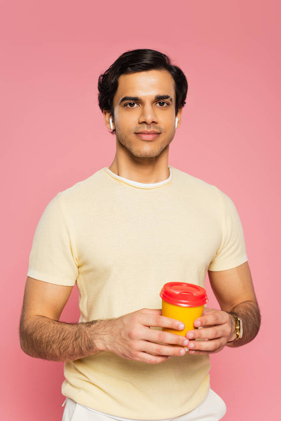 pleased young man in wireless earphones holding paper cup with coffee to go isolated on pink - Foto, imagen