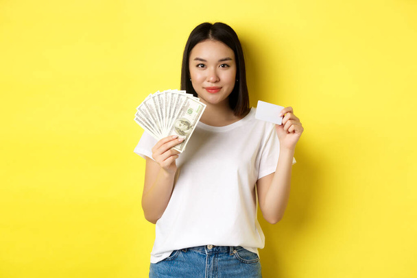 Kaunis aasialainen nainen lyhyet tummat hiukset, yllään valkoinen t-paita, näyttää rahaa dollareina ja muovinen luottokortti, seisoo keltainen tausta - Valokuva, kuva