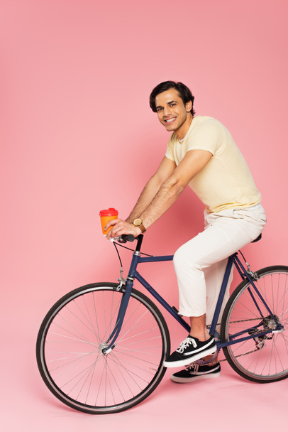 onnellinen nuori mies ratsastus polkupyörällä ja pitämällä paperimukia vaaleanpunaisella - Valokuva, kuva