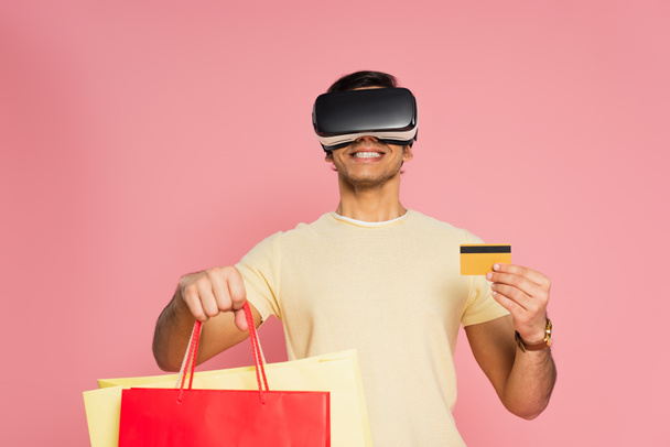 fröhlicher Mann in vr Headset mit Einkaufstaschen und Kreditkarte isoliert auf rosa - Foto, Bild