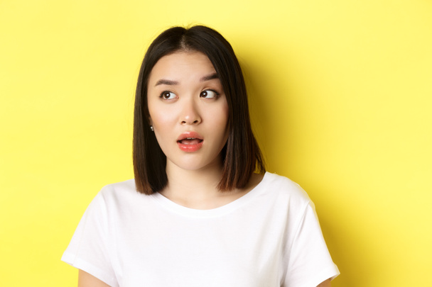 Close up of beautiful asian woman with clean perfect skin, raising eyebrow and looking left with intrigued face, standing over yellow background - Photo, Image
