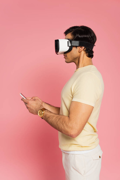boční pohled na mladého muže ve sluchátkách pomocí smartphonu na růžové - Fotografie, Obrázek