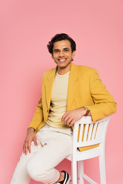cheerful young man in stylish blazer sitting on chair isolated on pink  - Photo, Image