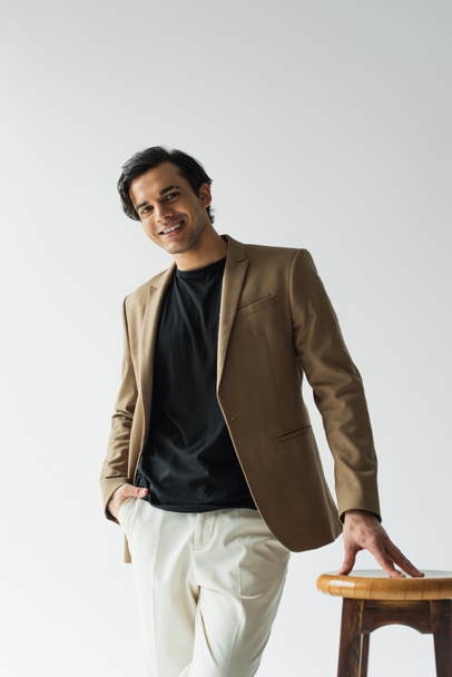 happy young man in stylish beige blazer posing with hand in pocket near wooden chair isolated on grey - Fotoğraf, Görsel