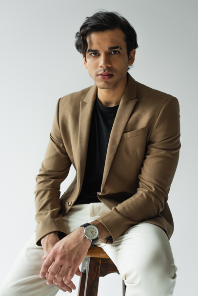 young man in stylish beige blazer looking at camera while sitting isolated on grey - Foto, Imagem