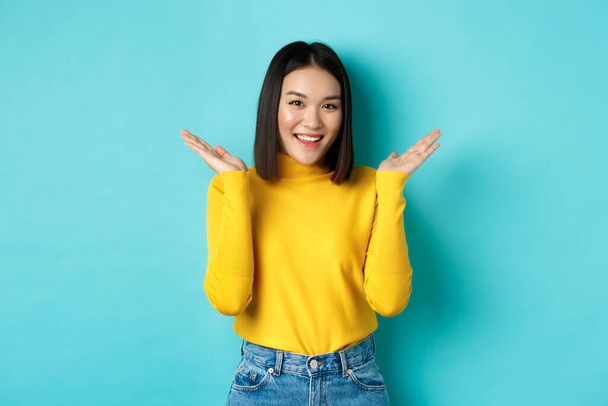 Piękno i moda. Atrakcyjny japoński dziewczyna podnieść ręce do góry i zademonstrować coś, uśmiechnięty szczęśliwy i patrząc w aparat fotograficzny, pokazując promo, niebieskie tło - Zdjęcie, obraz