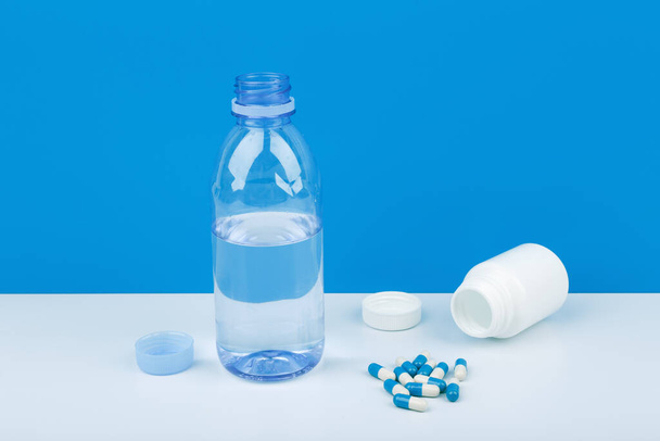 High angle view of opened transparent plastic bottle with water and medication bottle with spilled pills on white table against blue background. Concept of medical depression and bipolar treatment - Fotoğraf, Görsel