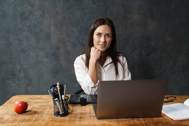 Smiling mid aged brunette businesswoman in shirt sitting at the desk with laptop computer over dark background - Φωτογραφία, εικόνα
