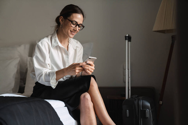 Smiling mid aged brunette white businesswoman sitting on bed in a hotel room holding mobile phone - Фото, зображення