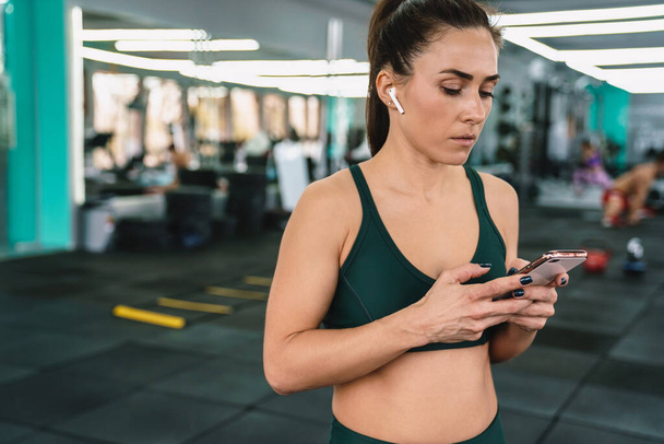 White sportswoman using mobile phone and earphones while working out in gym - Zdjęcie, obraz