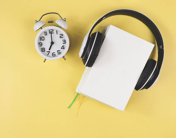 Top view or flat lay of white books  with headphones around and white vintage alarm clock shows 7 o'clock on yellow background. Audio books, morning routine concept. - Foto, Imagen