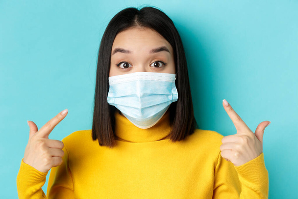 Covid-19, social distancing and pandemic concept. Close up of surprised asian girl pointing at her medical mask and looking amazed at camera, blue background - Photo, Image