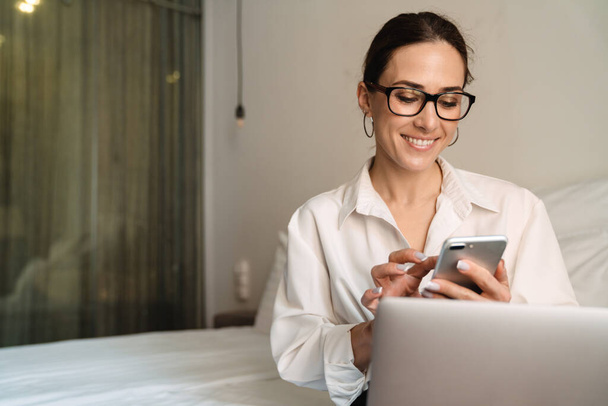 Smiling mid aged brunette white businesswoman sitting on bed in a hotel room typing on mobile phone working on laptop - Photo, image