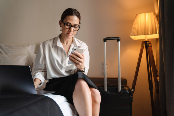 Mid aged brunette white businesswoman sitting on bed in a hotel room typing on mobile phone working on laptop - Fotoğraf, Görsel