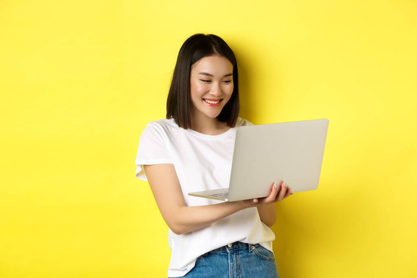 Cute asian female student working on laptop, reading screen and smiling, yellow background - Foto, immagini