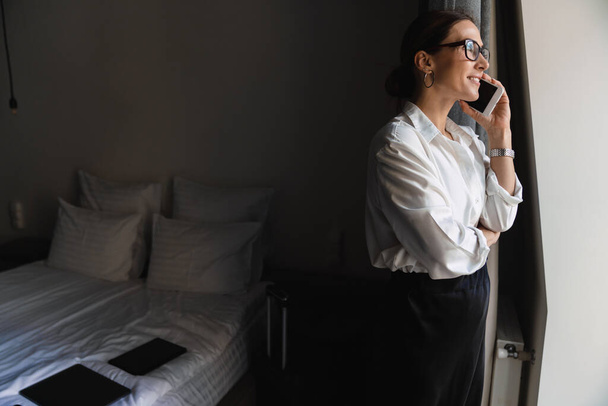 Χαμογελαστή μεσήλικη μελαχρινή λευκή επιχειρηματίας σε smart wear στέκεται στο παράθυρο του δωματίου του ξενοδοχείου μιλώντας στο κινητό τηλέφωνο - Φωτογραφία, εικόνα