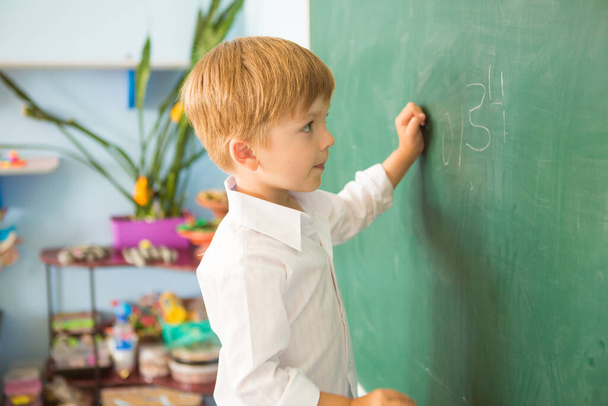 A little cute boy at school is standing near the blackboard and writing on it. Back to school concept - Foto, Imagen