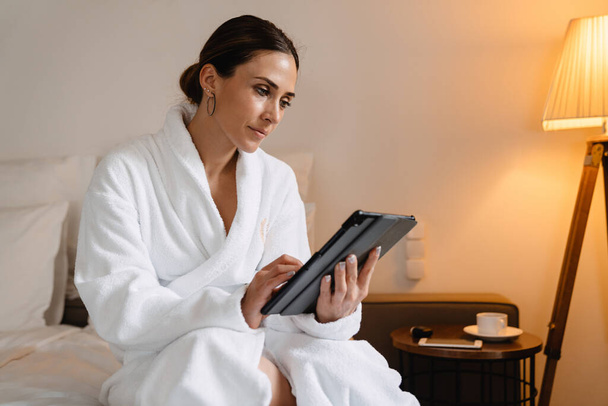 Smiling middle aged brunette white woman in bathrobe sitting on bed indoors holding tablet computer - Zdjęcie, obraz