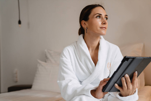 Smiling middle aged brunette white woman in bathrobe sitting on bed indoors holding tablet computer - Foto, afbeelding