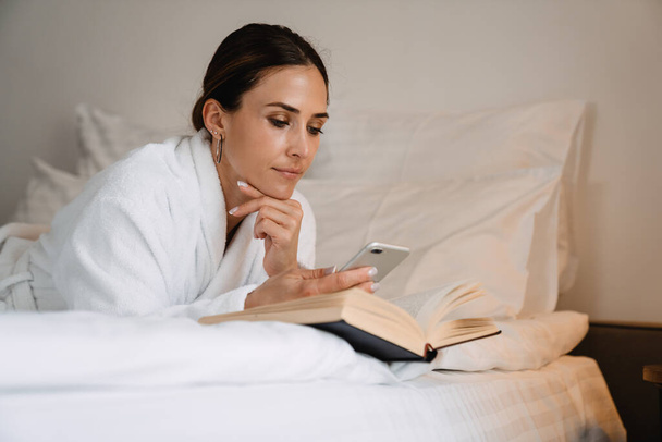 Smiling middle aged brunette white woman in bathrobe laying on bed indoors reading a book - Foto, Bild