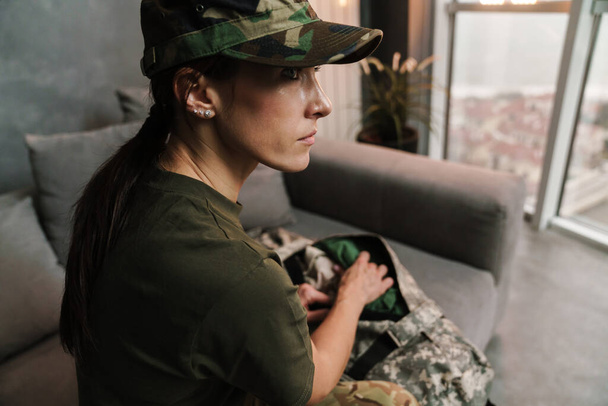 Serious beautiful soldier woman sitting on sofa with bag indoors - Фото, изображение