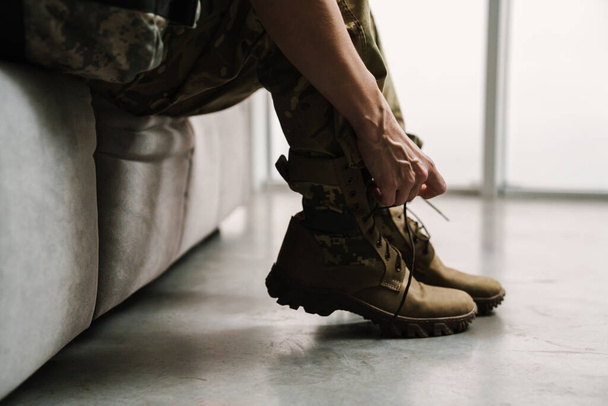 Soldier woman tying her shoe laces while sitting on couch indoors - Фото, изображение