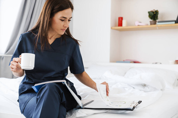 The brunette woman in blue medical uniform reads documents with a cup in her hands sitting on the bed in the room - Fotoğraf, Görsel