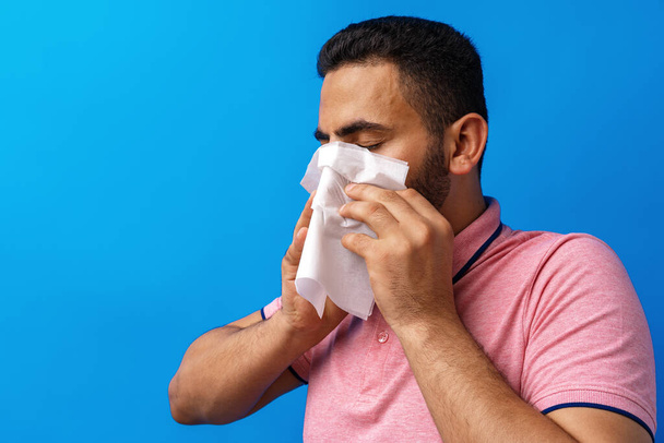 Young man in pink shirt with allergy or cold, blowing his nose in a tissue against blue background - Foto, Imagen