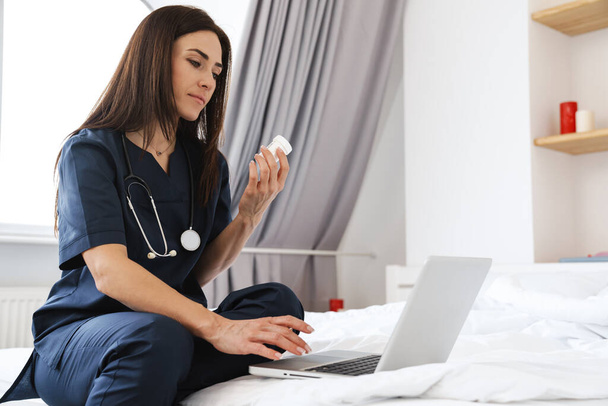 Focused woman doctor with stethoscope holding medicine while working with laptop at home - Foto, immagini