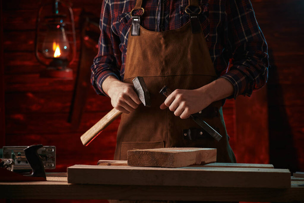 Ein Tischler im karierten Hemd und brauner Schürze hält einen Meißel in der Hand. - Foto, Bild
