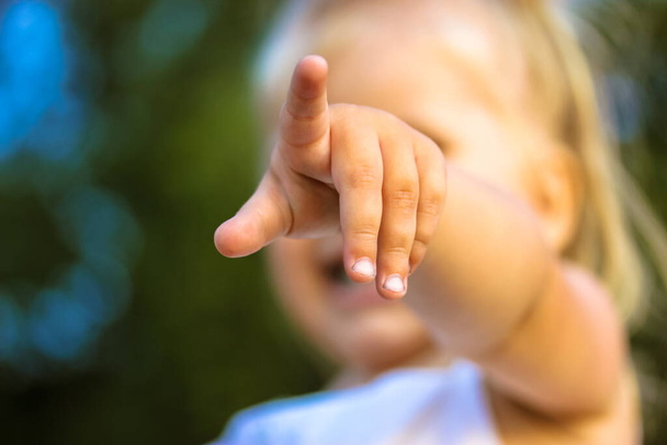 A little blonde infant girl pointing finger into a distance straight ahead. Baby girl's hand in focus. Age 2-3 years old. Young children in a green park blurry background at summer day. Cute child. - Foto, Imagem