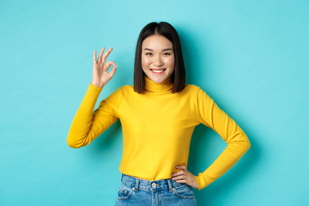 Boldog gyönyörű ázsiai nő mosolyog, mutatja rendben gesztus jóváhagyása, azt mondja, igen, állva elégedett a kék háttér - Fotó, kép