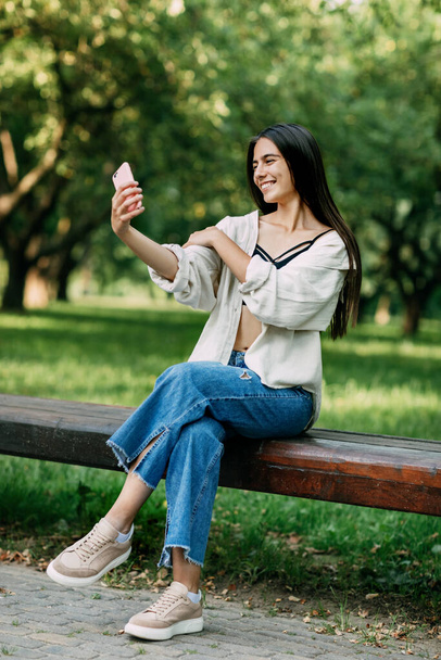 Atraktivní veselá brunetka s dlouhými vlasy emocionálně bere selfie sedí na lavičce v parku. Koncept relaxační dovolené - Fotografie, Obrázek