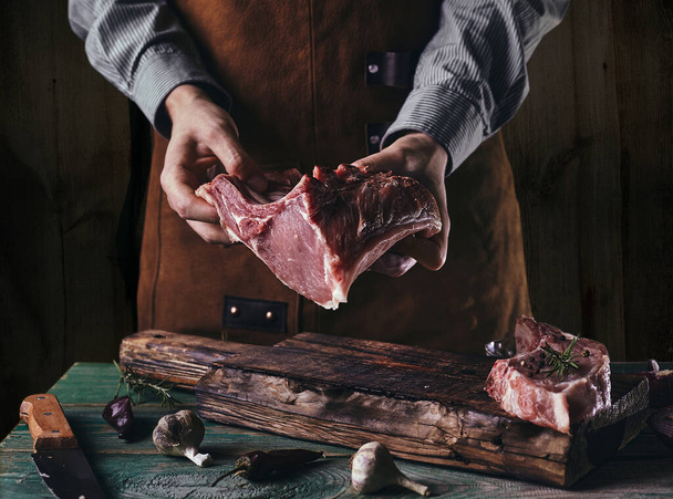 A man in a leather apron holds a piece of raw meat with a bone. Pork ribs in the hands of the butcher. - Zdjęcie, obraz