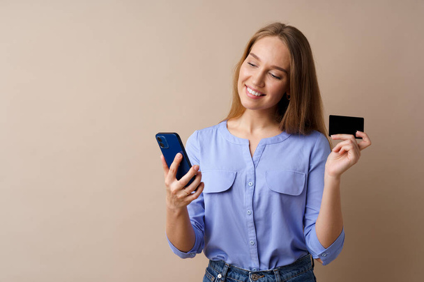 Happy young woman holding smartphone and credit card against beige background - Valokuva, kuva