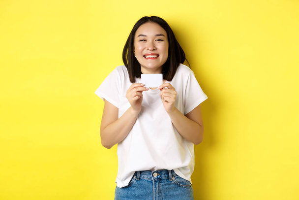Junge asiatische Frau in lässigem weißem T-Shirt geht einkaufen, zeigt Plastikkreditkarte und lächelt aufgeregt, steht über gelbem Hintergrund - Foto, Bild